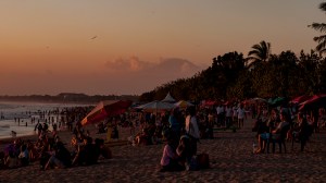 A volcano peeps out from the clouds as the crowds gather to watch sunset from Kuta Beach.
